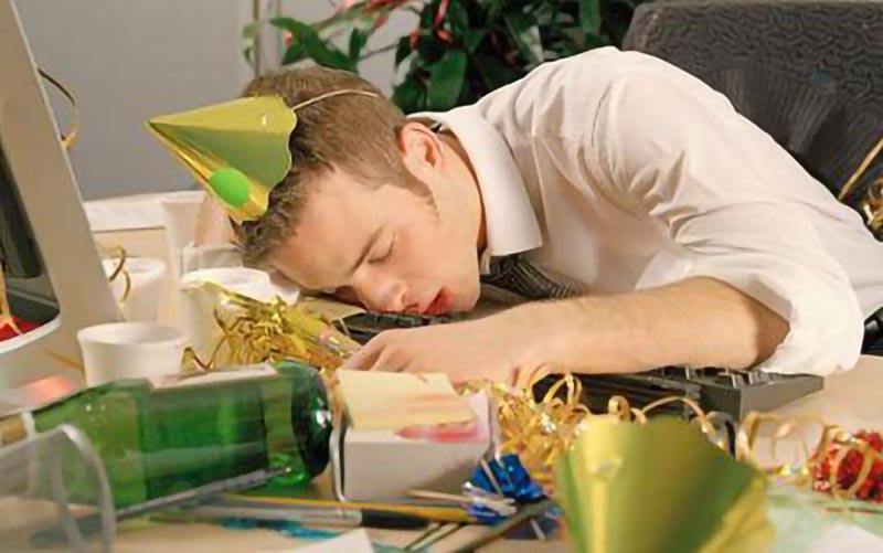 Drunk office worker wearing party hat asleep at computer desk with empty bottle